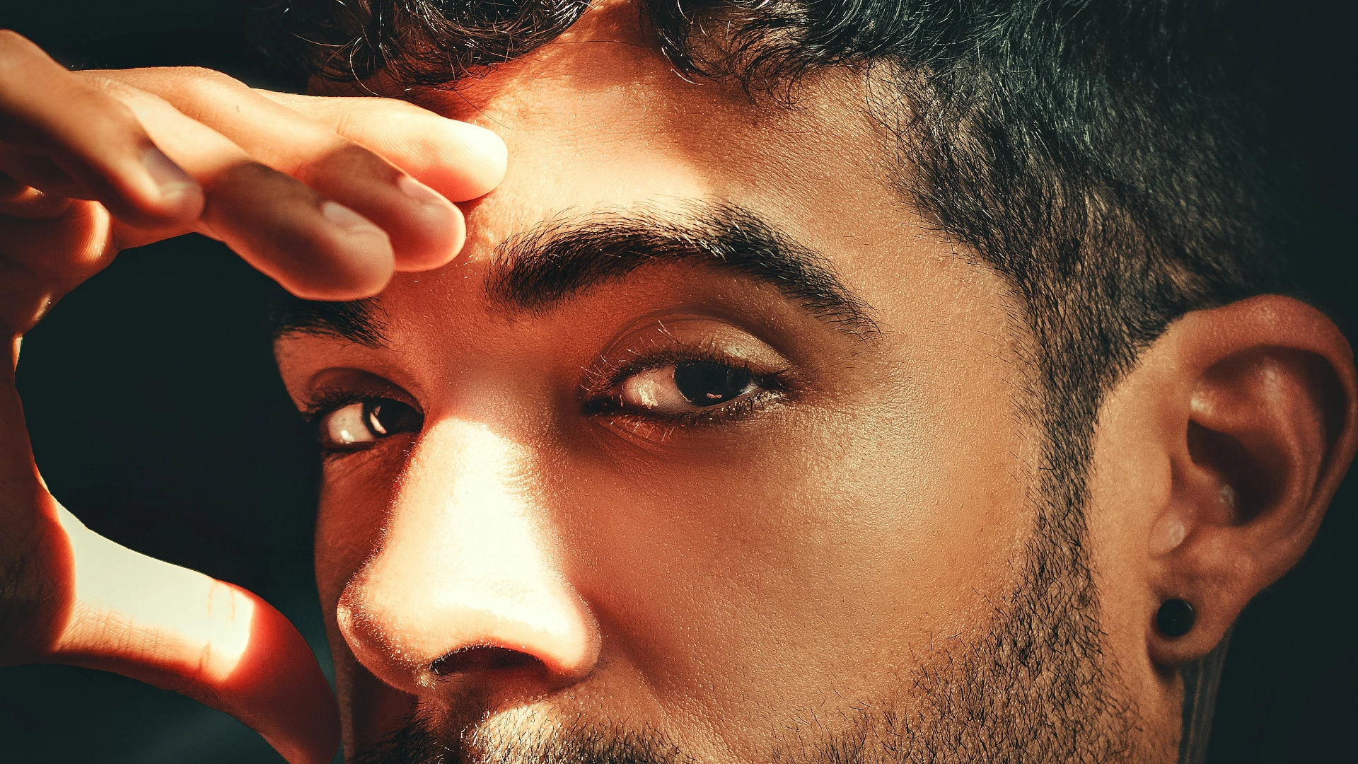 Close-up of a man with facial hair and earring resting his hand on his forehead
